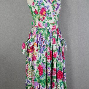 K&ouml;nnte beinhalten: Ein wei&szlig;es, &auml;rmelloses Kleid mit einem Blumendruck in Rosa, Lila und Gr&uuml;n. Das Kleid hat einen runden Ausschnitt, eine Knopfleiste vorne und einen gerafften Rock. Die Tr&auml;ger haben Schleifen an den Schultern.