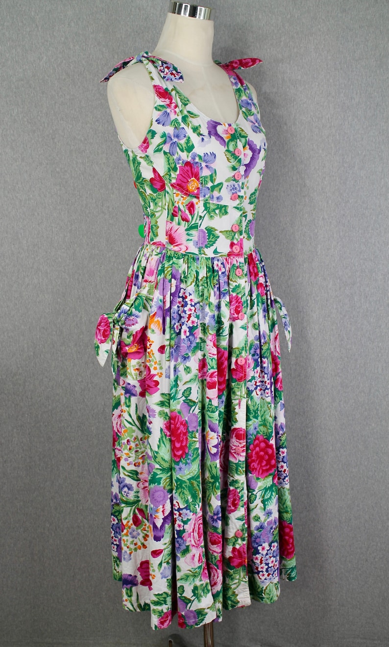K&ouml;nnte beinhalten: Ein wei&szlig;es, &auml;rmelloses Kleid mit einem Blumendruck in Rosa, Lila und Gr&uuml;n. Das Kleid hat einen runden Ausschnitt, eine Knopfleiste vorne und einen gerafften Rock. Die Tr&auml;ger haben Schleifen an den Schultern.