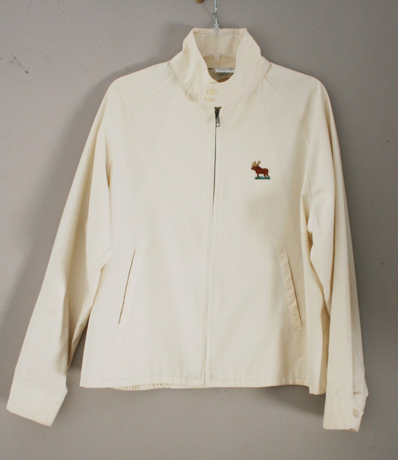 Puede incluir: Una chaqueta blanca con cremallera con un alce bordado en la parte delantera. La chaqueta tiene un cuello alto y dos bolsillos.