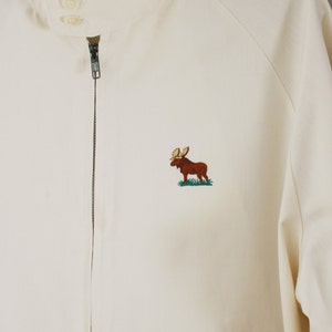 Puede incluir: Una chaqueta blanca con cremallera con un alce marr&oacute;n bordado en la parte delantera. La chaqueta tiene un cuello alto y una cremallera.