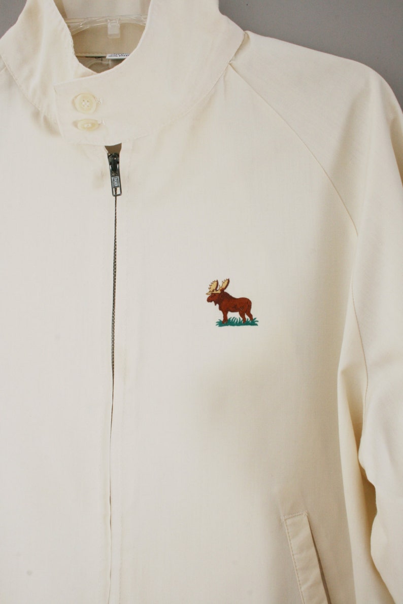 Puede incluir: Una chaqueta blanca con cremallera con un alce marr&oacute;n bordado en la parte delantera. La chaqueta tiene un cuello alto y una cremallera.