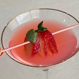 May include: A pink cocktail in a glass with a gold rim, garnished with a fresh strawberry on a light pink pick. The pick features a small, textured seashell at one end, resting across the rim of the glass, partially submerged in the liquid.