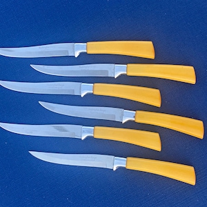 May include: A set of six steak knives with bright yellow handles and silver-colored metal blades. Each knife features a curved, pointed tip and a smooth, ergonomic handle design. The blades are made of stainless steel with a polished finish.