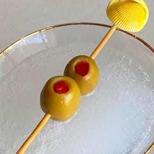 May include: A cocktail pick featuring a yellow seashell topper and two green olives stuffed with red pimentos, resting across a clear glass with a gold rim containing a translucent liquid with visible ice particles and bubbles.
