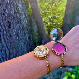 May include: A gold bracelet with a round, hinged compartment that opens to reveal a pink lip balm. The compartment is engraved with a floral design.