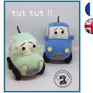 以下が含まれることがあります： 緑と青の2台の編みぐるみのおもちゃの車。黒い目と白い顔をしています。車は黒い縫い目の口と黒い縫い目の鼻をしています。車は白い表面の上に置かれています。車の真上に黒い文字で「tut tut!」と書かれています。