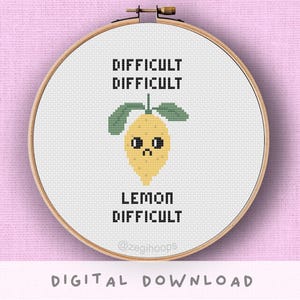 May include: A cross-stitch embroidery hoop with a yellow lemon design, sad face, and green leaves. The text reads "DIFFICULT DIFFICULT LEMON DIFFICULT". The embroidery is set against a white background and framed in a wooden hoop.
