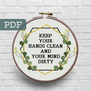 May include: A cross-stitch embroidery hoop with a white background and a gold geometric frame. The text inside the frame reads "Keep your hands clean and your mind dirty". The frame is surrounded by green eucalyptus leaves.