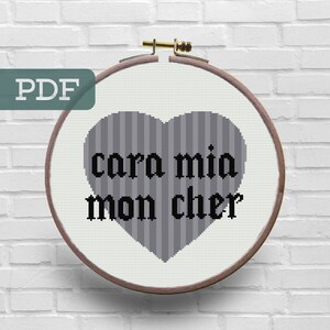 May include: A cross-stitch pattern featuring a gray heart with the text "cara mia mon cher" in black.