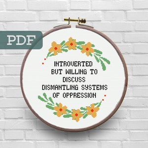 Puede incluir: Un aro de bordado blanco con un marco de madera marrón. El aro contiene un diseño de punto de cruz con un borde floral y el texto "Introvertido pero dispuesto a discutir el desmantelamiento de los sistemas de opresión".