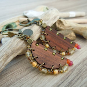 Puede incluir: Un par de pendientes de madera con forma de media luna, decorados con cuentas doradas y ámbar. Los pendientes están hechos de madera oscura y tienen un estilo rústico y bohemio.