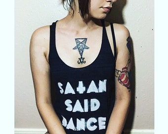 May include: A woman with black hair in a ponytail is wearing a black tank top with the text "Satan Said Dance" printed in white. She has a tattoo of a pentagram on her chest.
