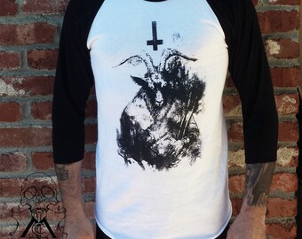 May include: A white baseball t-shirt with black sleeves features a black and white graphic print of a goat with an upside-down cross above its head.