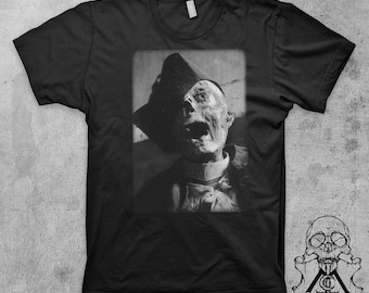 May include: Black t-shirt with a graphic print of a person's face with a hat on. The person's face is partially obscured by the hat. The person is wearing a white collar.
