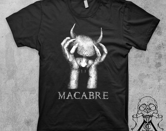 May include: Black t-shirt with a white graphic of a horned figure held by hands, with the word "MACABRE" below. A skull and crossbones design is on the lower right. The shirt is made of a soft material.