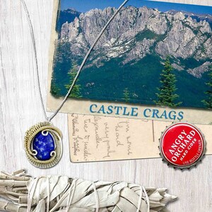 May include: A silver chain necklace with a wire-wrapped pendant featuring a blue lapis lazuli stone. The pendant is surrounded by gold wire. The necklace is displayed on a wooden surface with a postcard and a bottle cap.