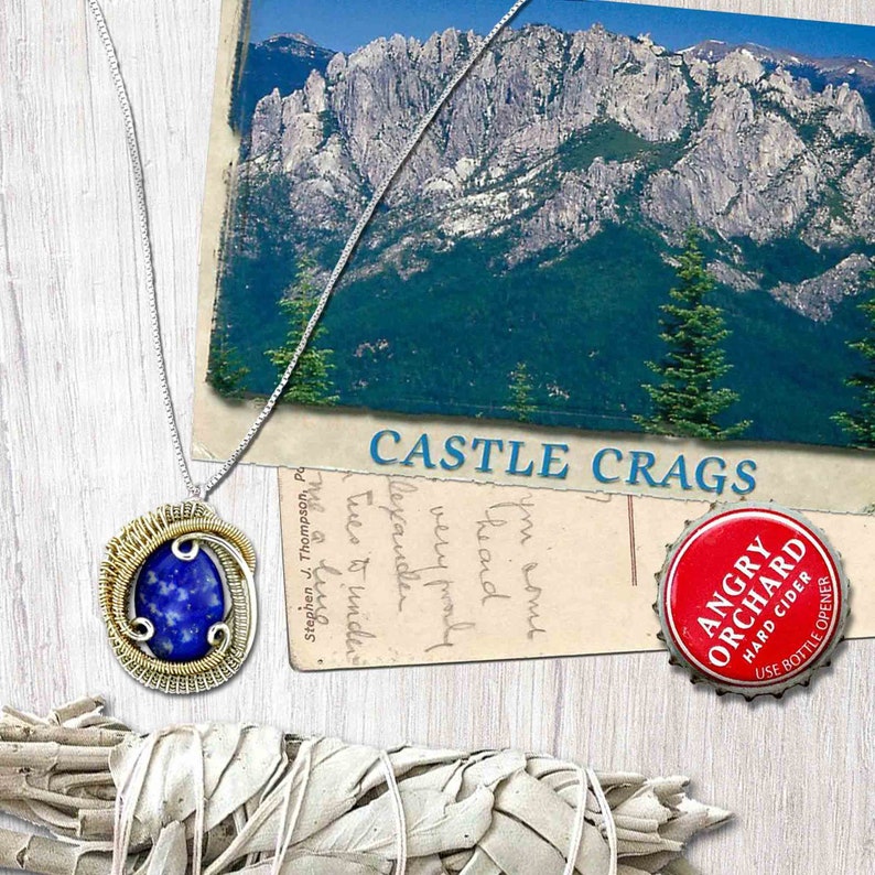 May include: A silver chain necklace with a wire-wrapped pendant featuring a blue lapis lazuli stone. The pendant is surrounded by gold wire. The necklace is displayed on a wooden surface with a postcard and a bottle cap.