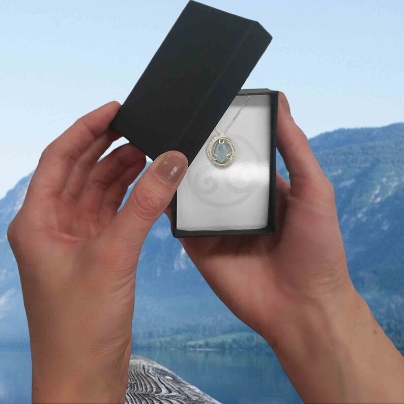 May include: A small, silver box with a blue and green gemstone pendant inside. The box is open and the pendant is visible. The background is a blurry image of a lake and mountains.