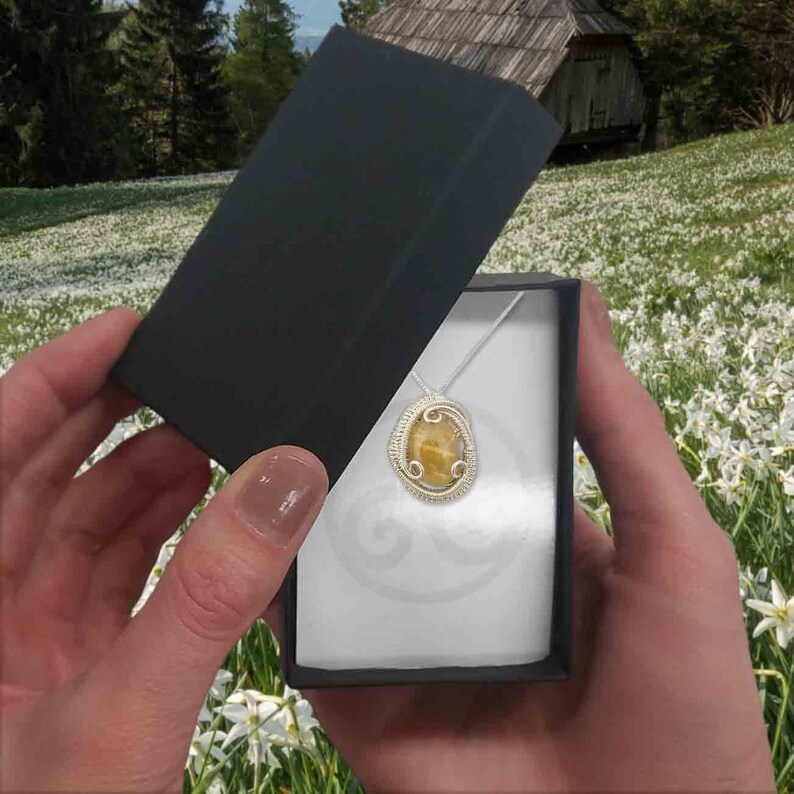 May include: A silver necklace with a yellow and brown oval pendant in a black and white gift box. The pendant is set in a silver wire frame.