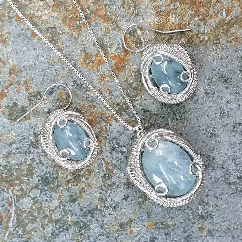 May include: A silver wire wrapped aquamarine pendant necklace and matching earrings set. The aquamarine stones are oval shaped and have a light blue color.