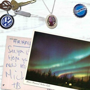 May include: A postcard featuring a photograph of the aurora borealis, or Northern Lights, above a dark forest. The postcard has handwritten text that reads "BORA-BORA AIR MAIL See you soon Hope you are well new year Mich +B Alaska's Phenomenal Northern Lights".