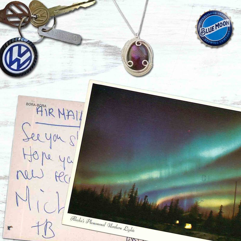 May include: A postcard featuring a photograph of the aurora borealis, or Northern Lights, above a dark forest. The postcard has handwritten text that reads "BORA-BORA AIR MAIL See you soon Hope you are well new year Mich +B Alaska's Phenomenal Northern Lights".