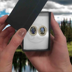 May include: A pair of silver wire wrapped earrings with oval-shaped blue and green iridescent stones. The earrings are displayed in a white box with a black lid.