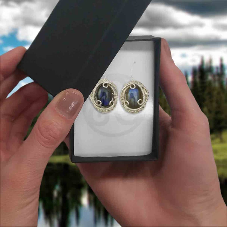 May include: A pair of silver wire wrapped earrings with oval-shaped blue and green iridescent stones. The earrings are displayed in a white box with a black lid.