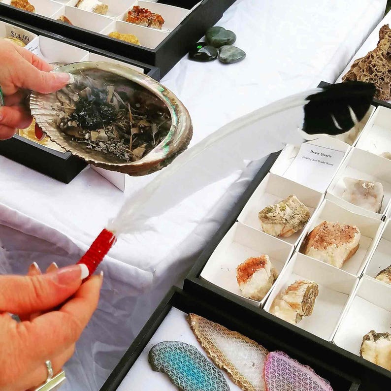 May include: A white feather with a black tip is being used to smudge a seashell with burning sage. The seashell is on a black tray with a price tag that reads &pound;14.99.  There are several small boxes filled with crystals and stones in the background.