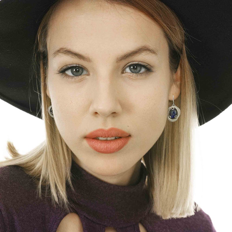 May include: A woman with blonde hair wearing a black hat and a purple turtleneck sweater. She is wearing earrings with blue stones.