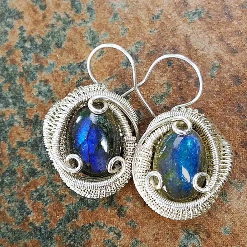May include: A pair of silver wire wrapped earrings with oval shaped labradorite gemstones. The gemstones have a blue and green iridescent sheen.