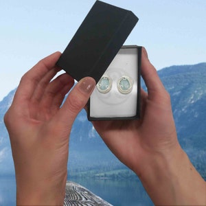 May include: A pair of silver earrings with light blue oval-shaped stones set in a delicate wire design. The earrings are displayed in a black gift box.