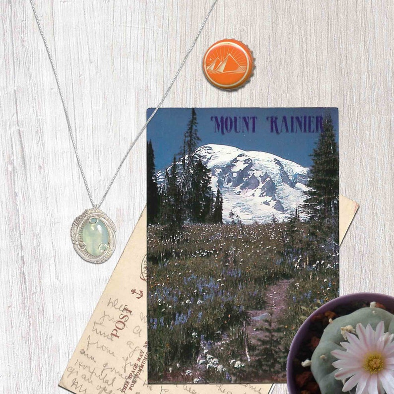 May include: A postcard featuring a scenic view of Mount Rainier, a snow-capped mountain with a lush green valley in the foreground. The postcard has the text "MOUNT RAINIER" printed in blue letters at the top.