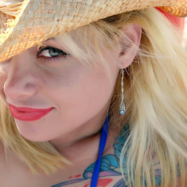 May include: A close-up of a woman's face with blonde hair, wearing a straw hat and a blue halter top. She is wearing a silver dangle earring with a blue gemstone.