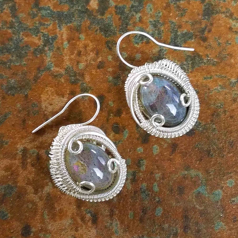 May include: A pair of silver wire wrapped earrings with iridescent labradorite gemstones. The earrings have a teardrop shape and are accented with silver wire.