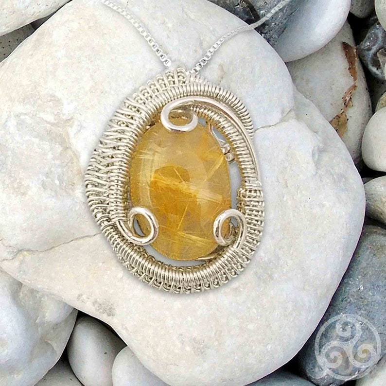 May include: A silver wire wrapped pendant featuring a golden rutilated quartz gemstone. The pendant is displayed on a bed of white stones.