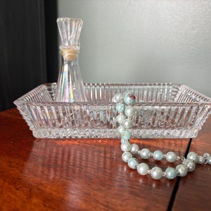 May include: A clear glass rectangular tray with a vertical ribbed pattern, containing a small clear glass bottle with a tapered neck and a string of light blue and white spherical beads with a subtle floral pattern on some of the beads.