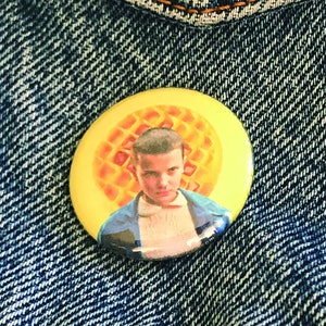 May include: A yellow button with a picture of a character from the show Stranger Things. The character is wearing a blue and white shirt and has brown hair. The button has a waffle pattern around the character's head. The text "www.bryanchuckbrennan.com" is printed around the edge of the button. The text "Bryan Chuck Brennan GROOVY-BUTTONS" is printed below the button.