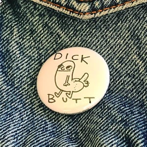 Puede incluir: Un pin blanco con las palabras "DICK BUTT" y una ilustración de dibujos animados de una figura con el trasero levantado. El pin está sobre una chaqueta vaquera azul. La dirección del sitio web "WWW.BRYANCHUCKBRENNAN.COM" es visible.
