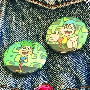 May include: Two round button pins featuring cartoon characters with green and blue hair. The characters are on a green and white background with a geometric pattern. The buttons are pinned to a blue denim jacket.
