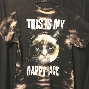 Puede incluir: Camiseta negra con un patr&oacute;n de tie-dye blanco. La camiseta presenta una imagen de un gato gru&ntilde;&oacute;n con el texto "This is my happy face".