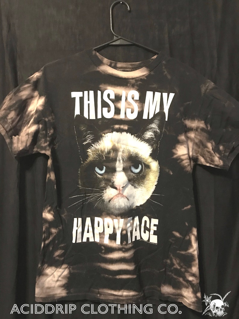 Puede incluir: Camiseta negra con un patr&oacute;n de tie-dye blanco. La camiseta presenta una imagen de un gato gru&ntilde;&oacute;n con el texto "This is my happy face".