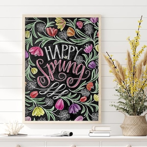 May include: A framed art print with the words "Happy Spring" in white and pink script, surrounded by colourful tulips and green foliage on a black background. The print is displayed above a white cabinet with woven baskets.