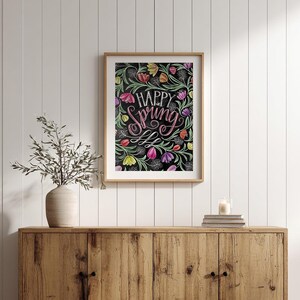 May include: A framed art print with the words "Happy Spring" in pink script, surrounded by colourful flowers on a black background. The artwork is in a wooden frame and displayed on a wooden cabinet.