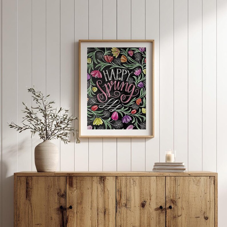May include: A framed art print with the words "Happy Spring" in pink script, surrounded by colourful flowers on a black background. The artwork is in a wooden frame and displayed on a wooden cabinet.