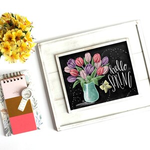 Puede incluir: Un cuadro de pizarra enmarcado con un ramo de tulipanes rosas y morados en un jarr&oacute;n turquesa, con las palabras "hello SPRING" en escritura blanca. Un peque&ntilde;o bloc de notas, un reloj y flores amarillas tambi&eacute;n est&aacute;n en la imagen.