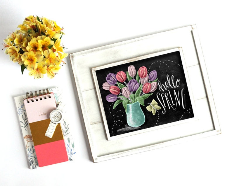 Puede incluir: Un cuadro de pizarra enmarcado con un ramo de tulipanes rosas y morados en un jarr&oacute;n turquesa, con las palabras "hello SPRING" en escritura blanca. Un peque&ntilde;o bloc de notas, un reloj y flores amarillas tambi&eacute;n est&aacute;n en la imagen.