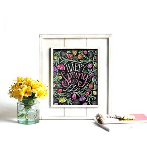 May include: A framed art print with the words "Happy Spring" in a cursive font, surrounded by colourful floral illustrations. The print is set against a black background and framed in a white wooden frame. A small vase of yellow flowers sits to the left.
