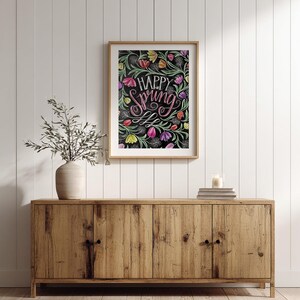 May include: A framed art print featuring the words "Happy Spring" in pink script, set against a backdrop of colourful floral illustrations on black. The artwork hangs above a wooden cabinet, with a vase and candle.