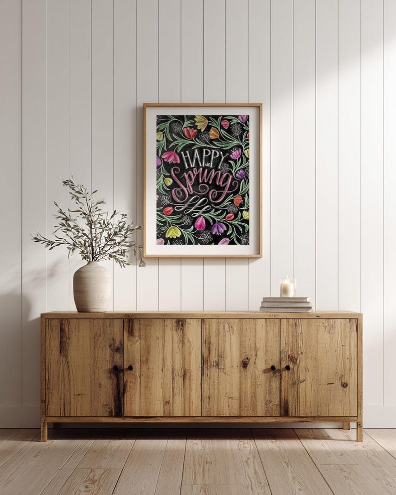 May include: A framed art print featuring the words "Happy Spring" in pink script, set against a backdrop of colourful floral illustrations on black. The artwork hangs above a wooden cabinet, with a vase and candle.